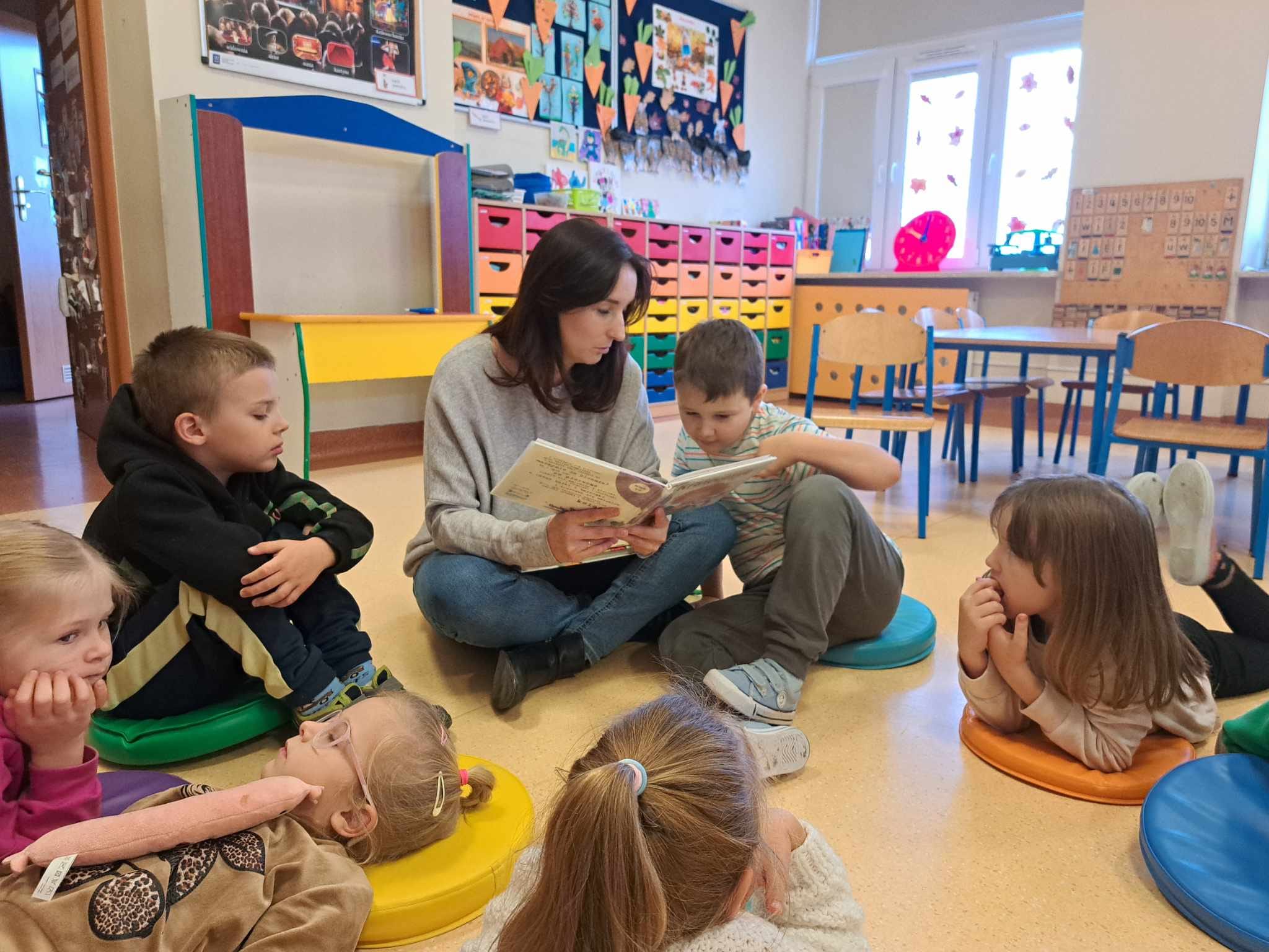 Dzieci siedzą na podłodze, słuchają tekstu czytanego przez kobietę