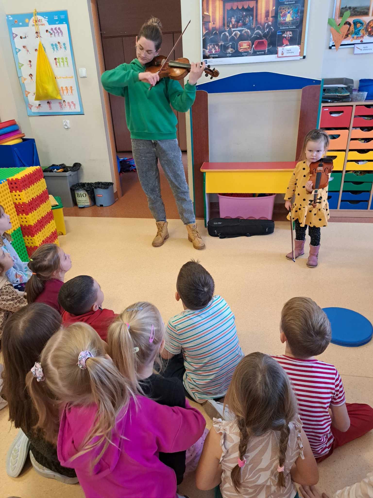 Dzieci siedzą na podłodze, słuchają osoby grającej na skrzypcach