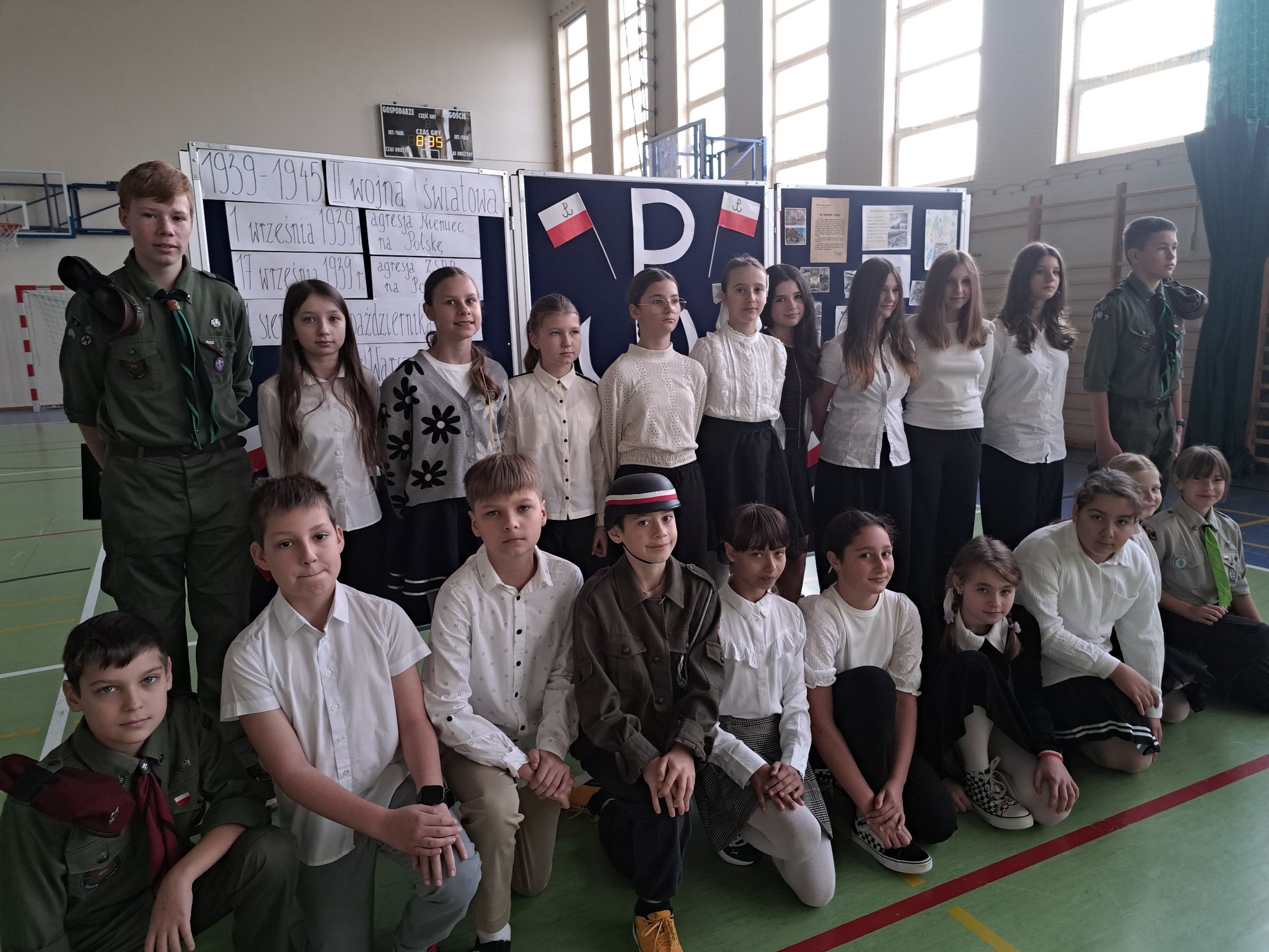 Uczniowie recytujący wiersze na tle dekoracji o powstaniu warszawskim